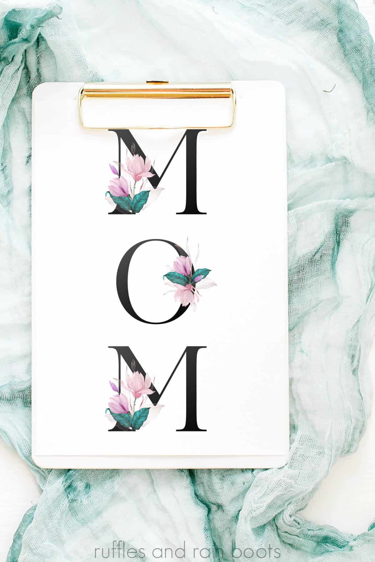 Vertical image of a white card on a small white clipboard on a scarf background with big, bold letters which spell out mom which have pink and green floral accents.