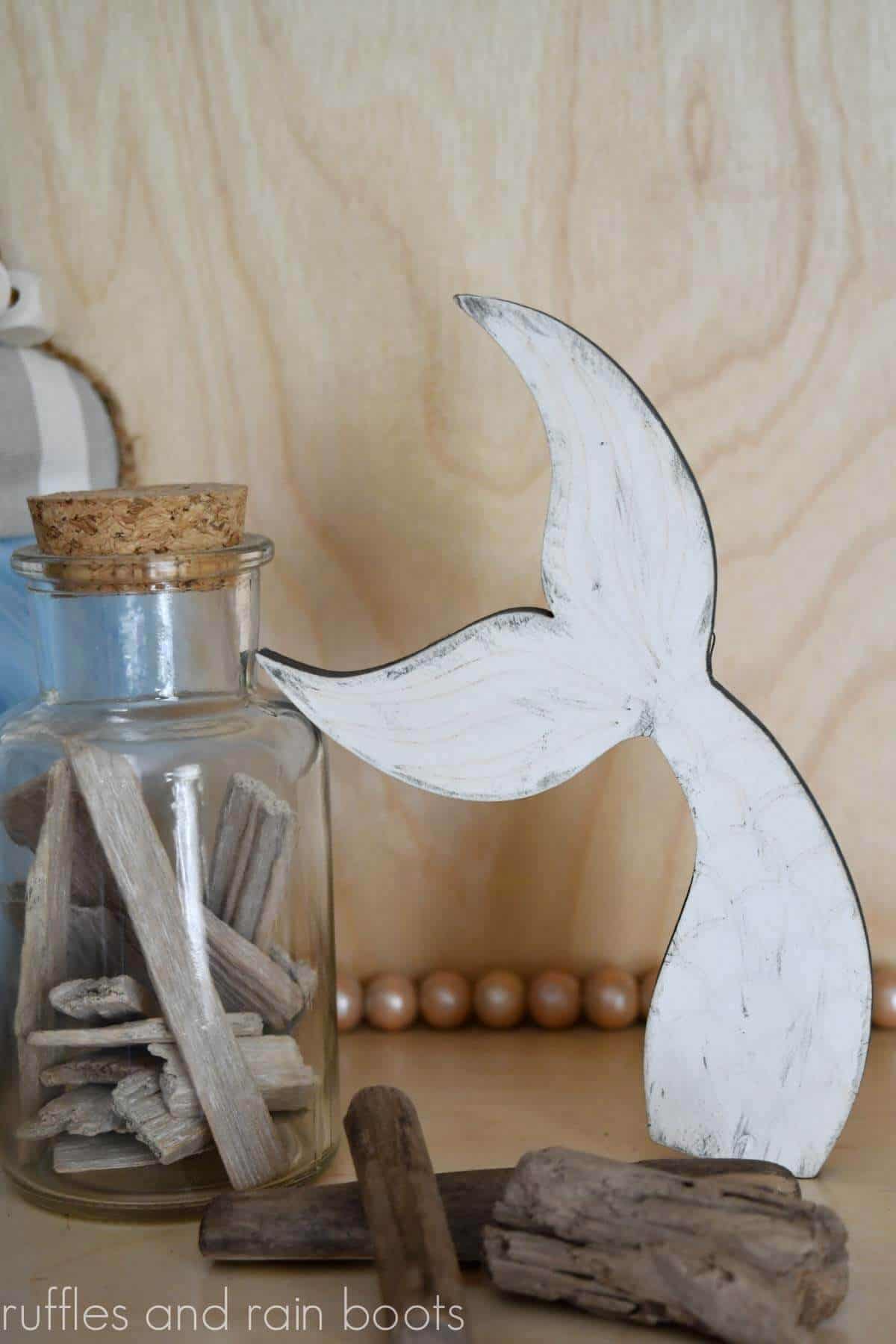 Vertical image of a mermaid tail shelf sitter on a light wood background with driftwood pieces scattered and in a glass jar with cork top.