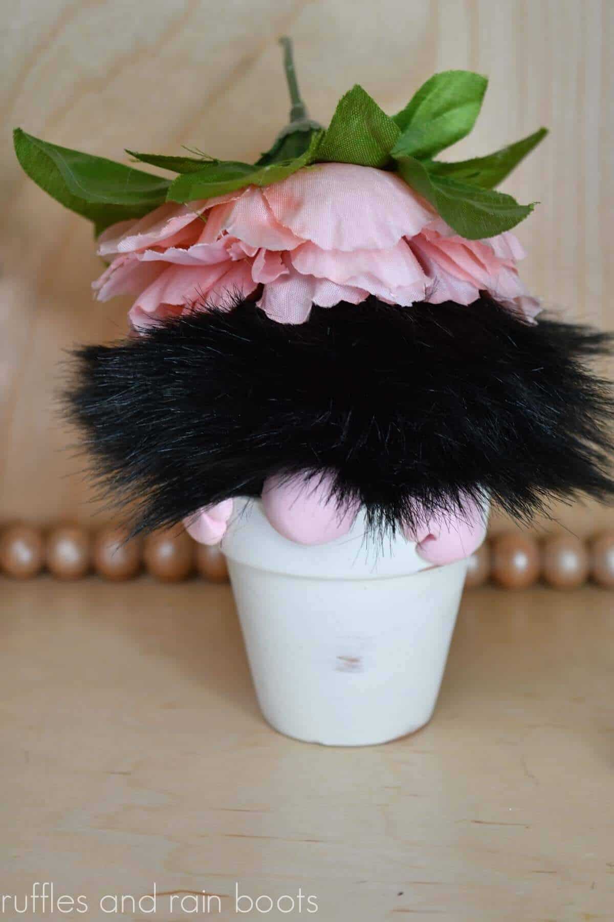 Vertical close up image of a black pompom gnome with a flower hat in a white miniature clay flower pot.