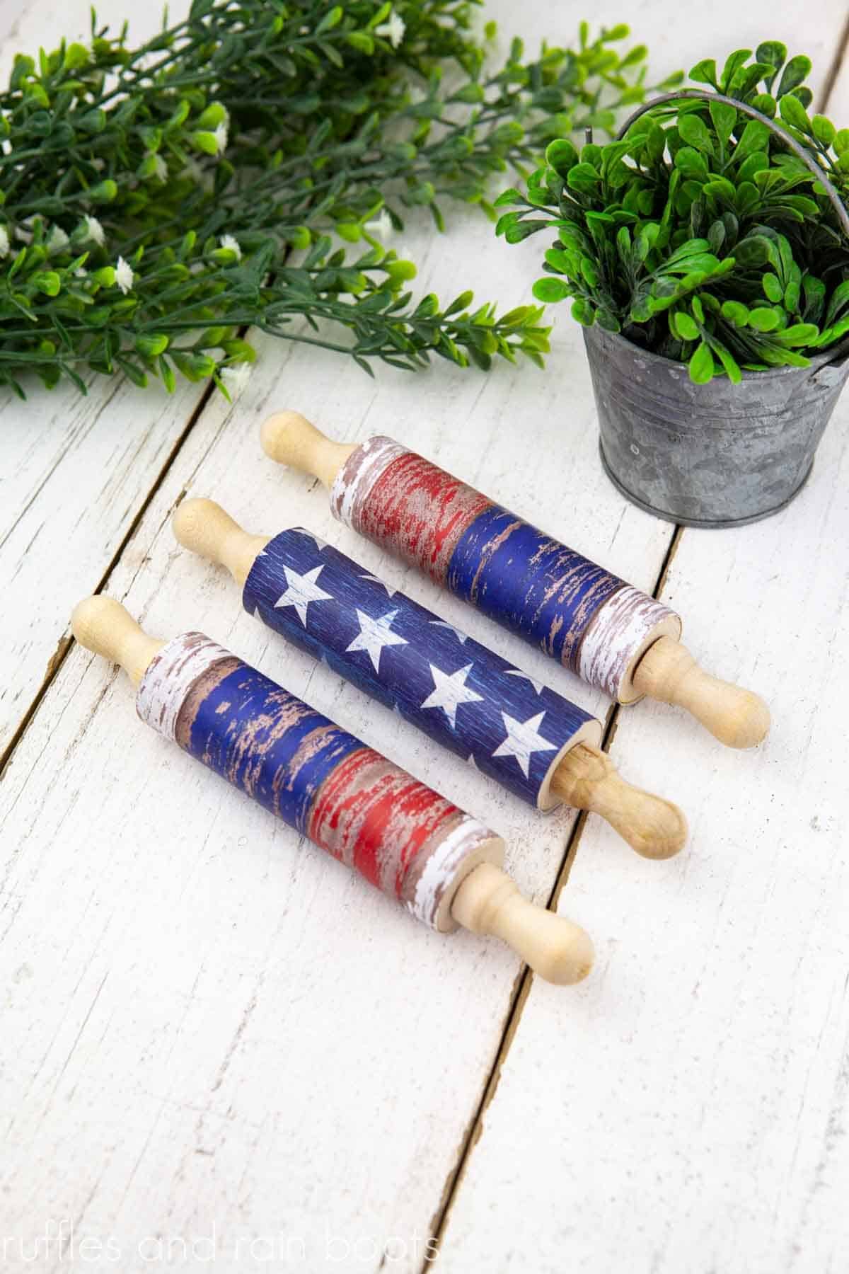 Vertical image of three paper covered farmhouse rolling pins for Independence Day decor on white wood with boxwood greenery.