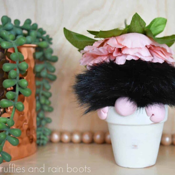 Square image of a mini gnome in a flower pot made with a black pompom and clay next to a copper vase with succulents.