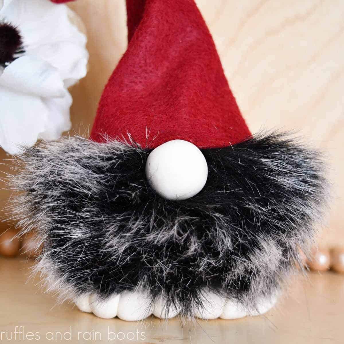 Square close up image of a black and white fur pompom gnome with clay feet, nose, and a red, twisty felt hat.