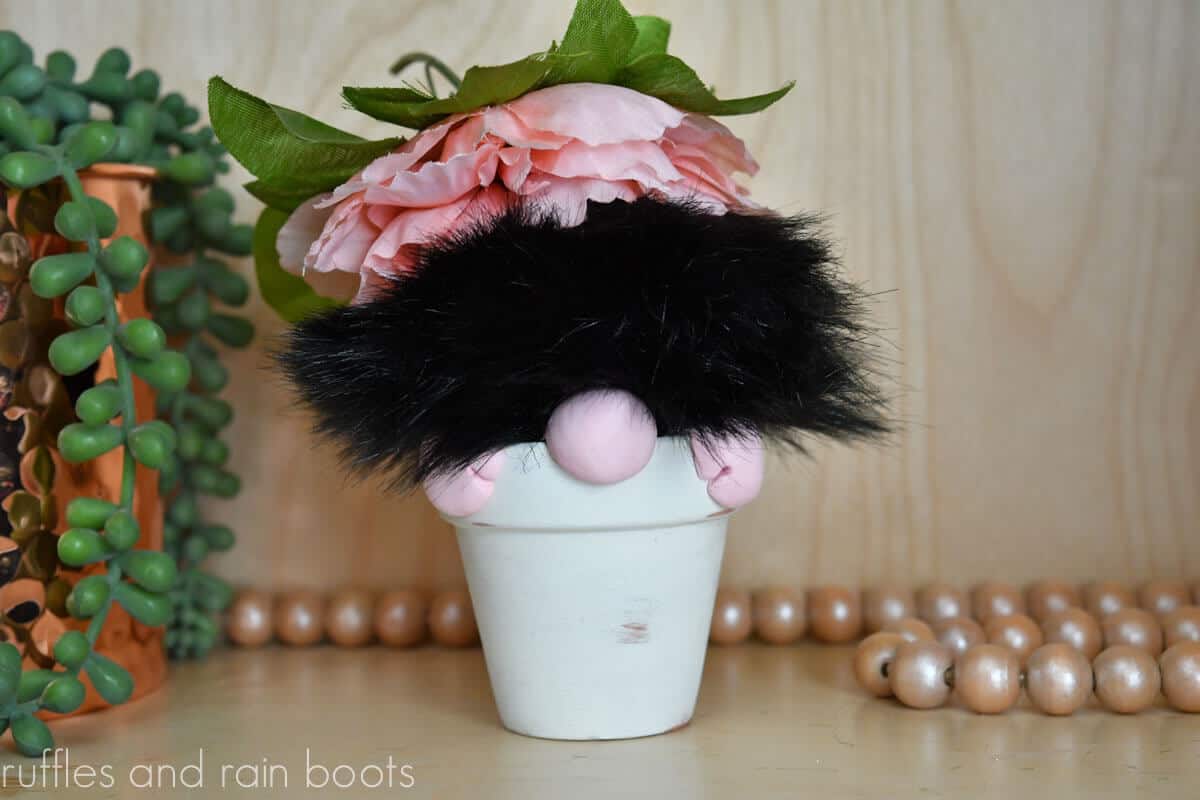 Horizontal image of a close up of a black faux fur pompom gnome in a white farmhouse clay pot planter in front of a light wood background.