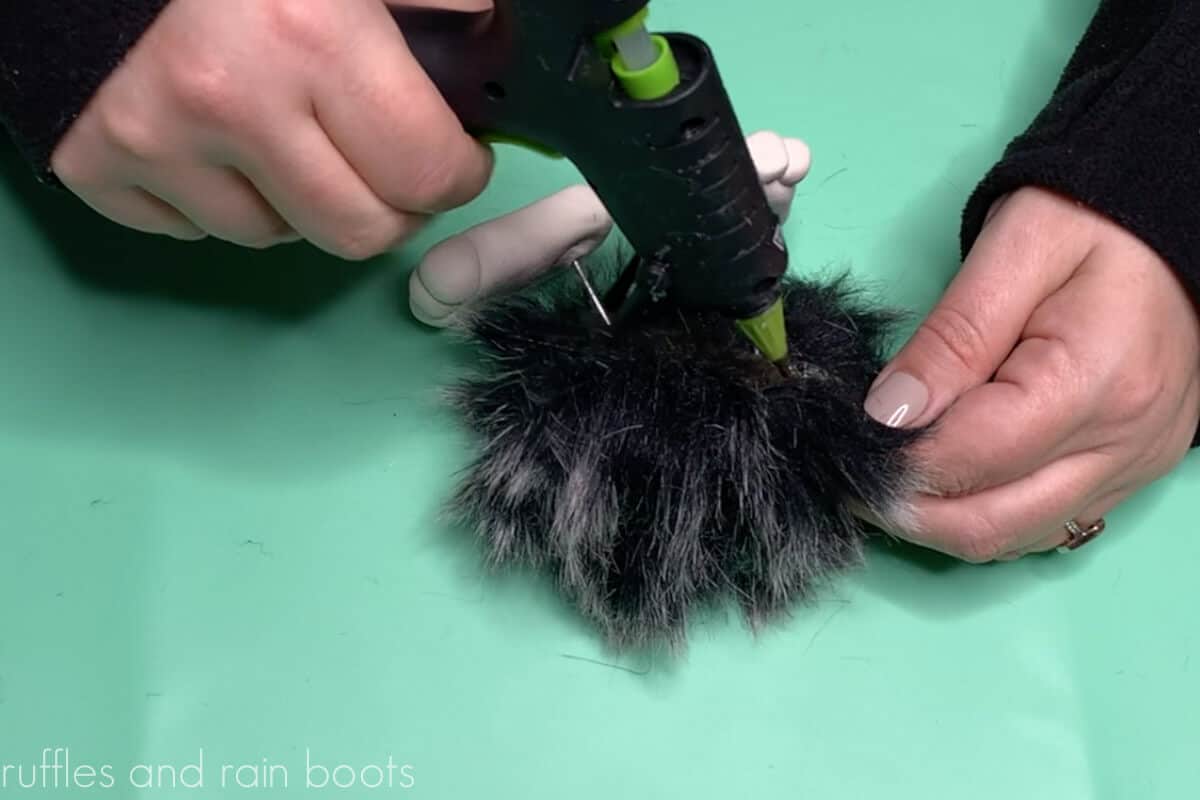 Crafter hands using a detail tip glue gun to close the fur pompom gnome body with legs inside.