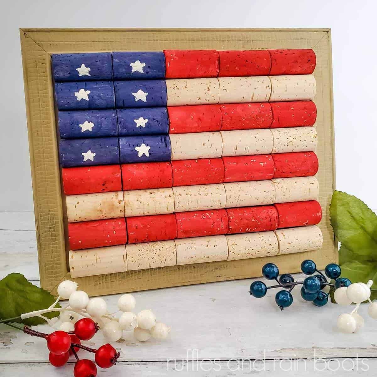 Square image close up of a wine cork flag painted in red and blue acrylic paint with small stars on a white wood background with red, white, and blue berries.
