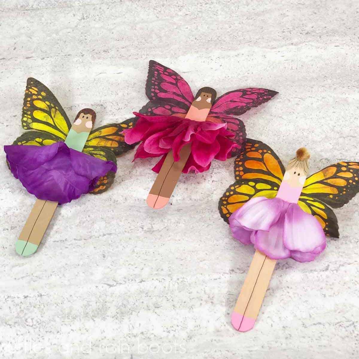 Square close up image of three craft stick fairies with flower skirts and butterfly wings on a light concrete background.