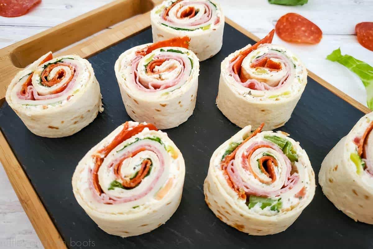 Italian meat pinwheel sandwiches on a black and wood cutting board on white wood background.