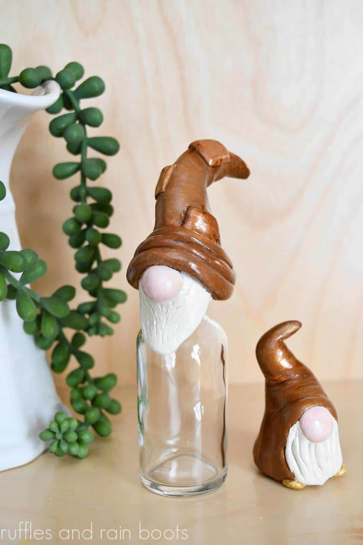 Vertical close up image of a clay gnome jar topper and a mini Model Magic gnome in front of a light wood background.