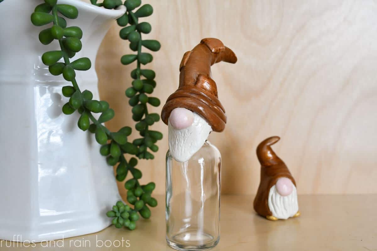 Horizontal image close up of a gnome jar topper in front of a light wood background standing next to a white vase with succulent.