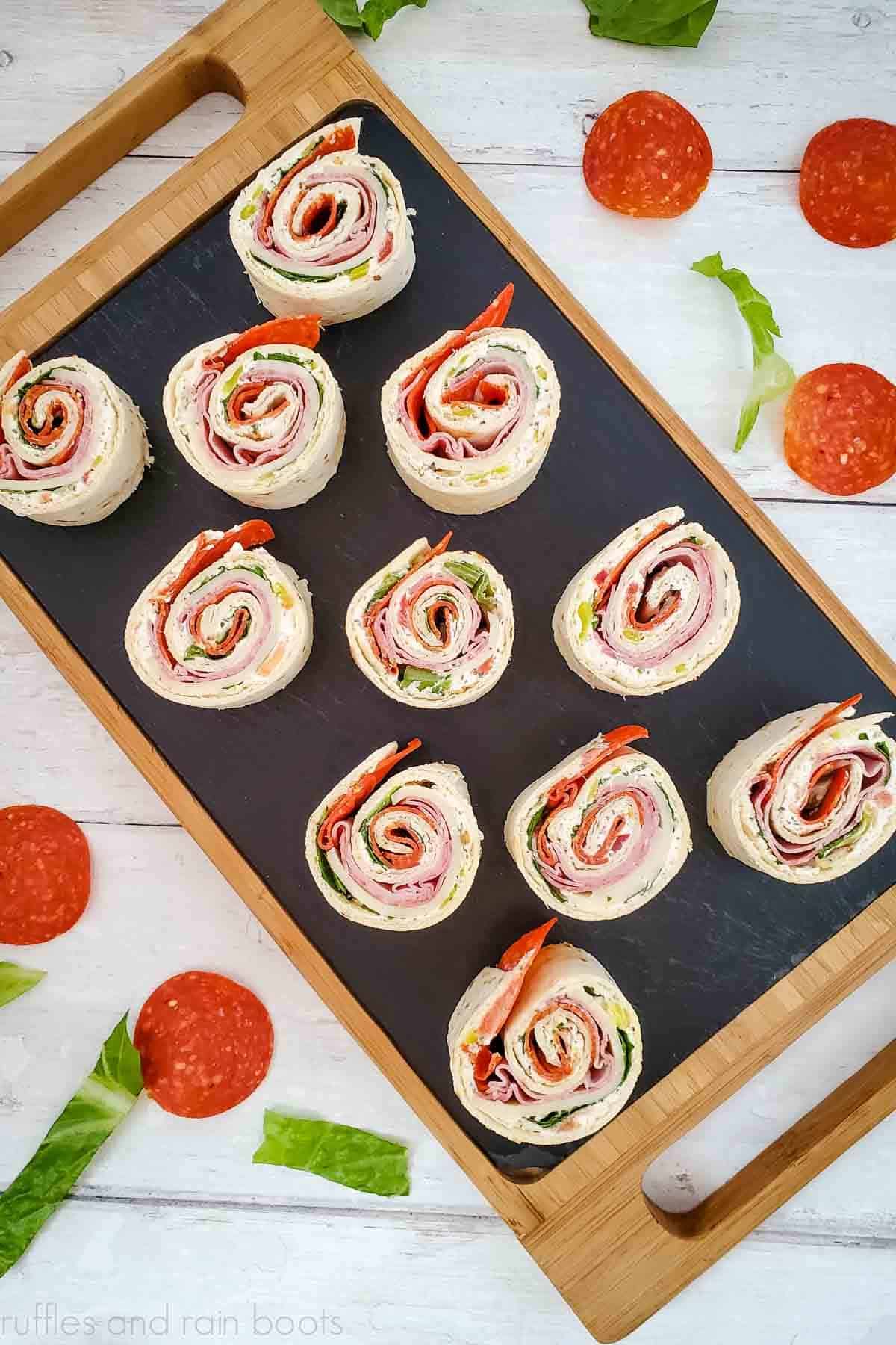 Vertical image of 10 Italian pinwheel sandwiches on a cutting board with pepperoni and lettuce on white wood.