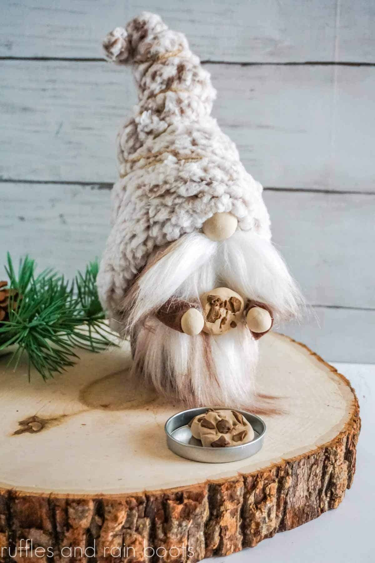 Vertical image of a gnome with a cookie on a wood log with pine branch in front of a white wood background.