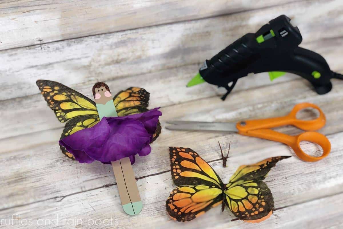 Horizontal image showing a hot glue gun attaching a butterfly to the back of a popsicle stick fairy on a light wood background.