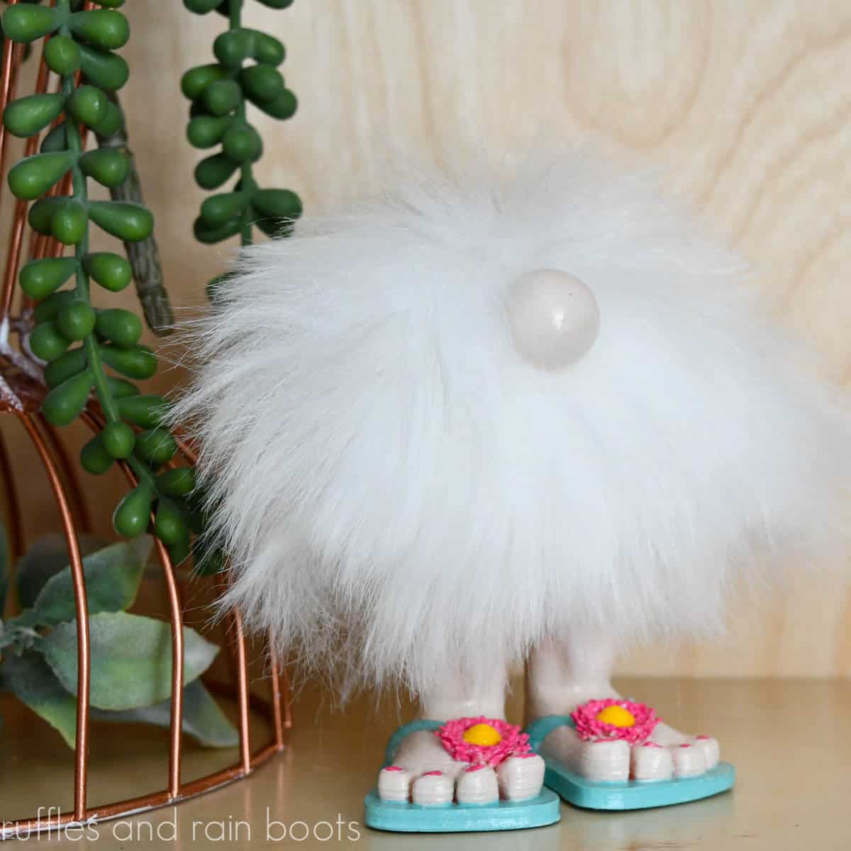 Square image close up of a white pompom gnome in pink and blue flip flops next to a plant in front of a light wood wall.