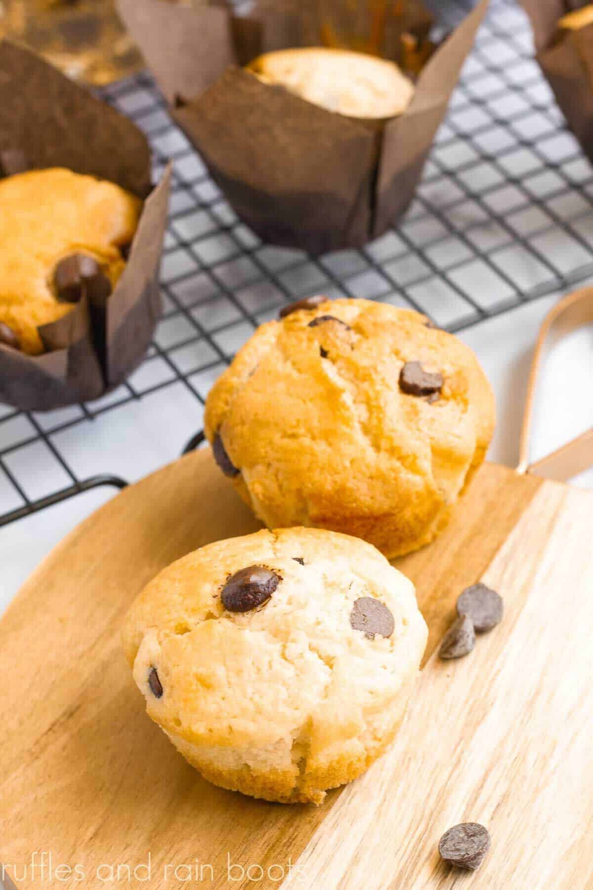 Vertical image of 2 chocolate chip muffins on a wood cutting board in front of muffins in paper wrappers on wire rack.