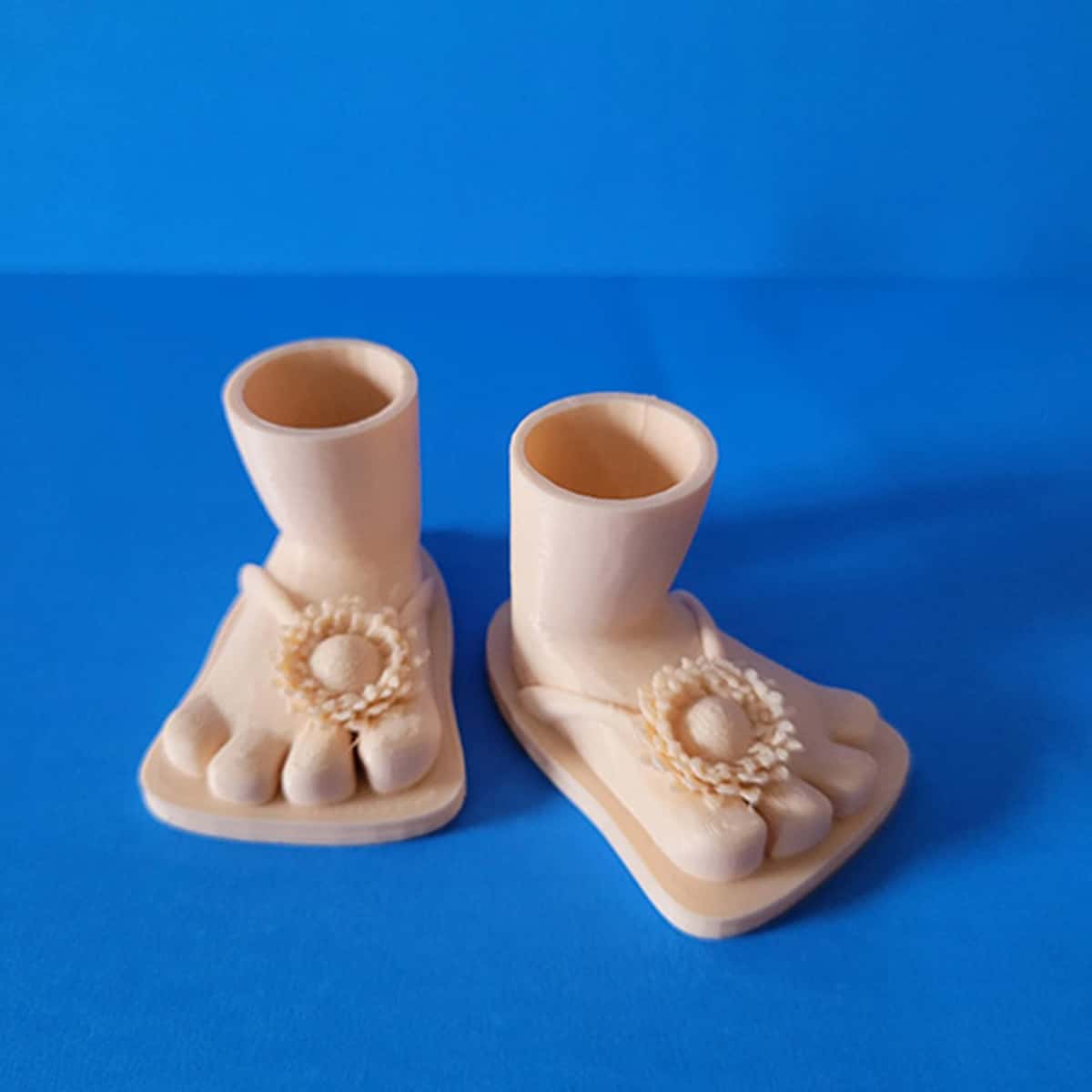 Square image of 3D printed gnome bare feet flip flop with flower on blue background.