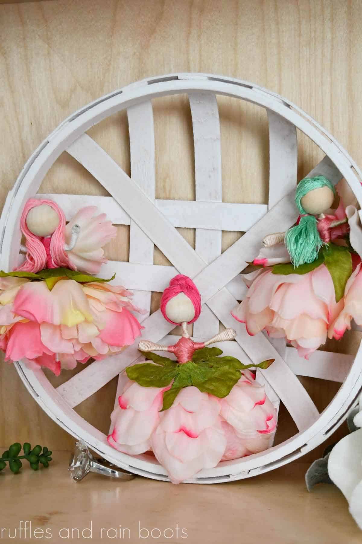 Vertical image of three flower fairy dolls with wood bead heads, floss hair, and silk flower skirts on white basket.