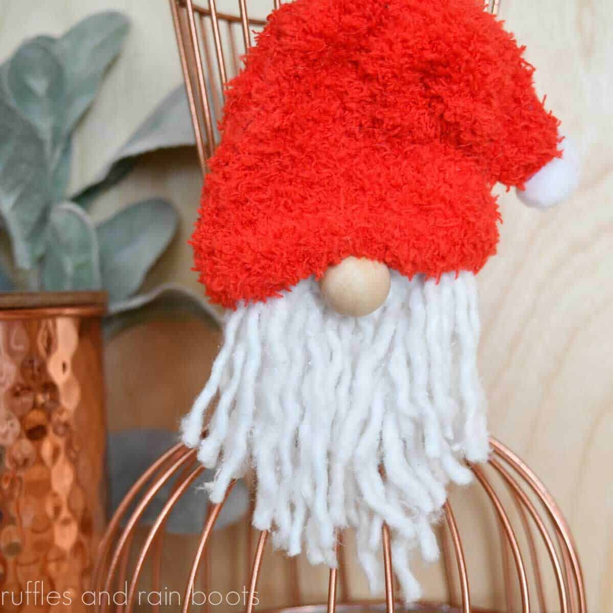 Red hat gnome ornament made from a curtain ring hanging on copper vase with greenery in front of light wood background.