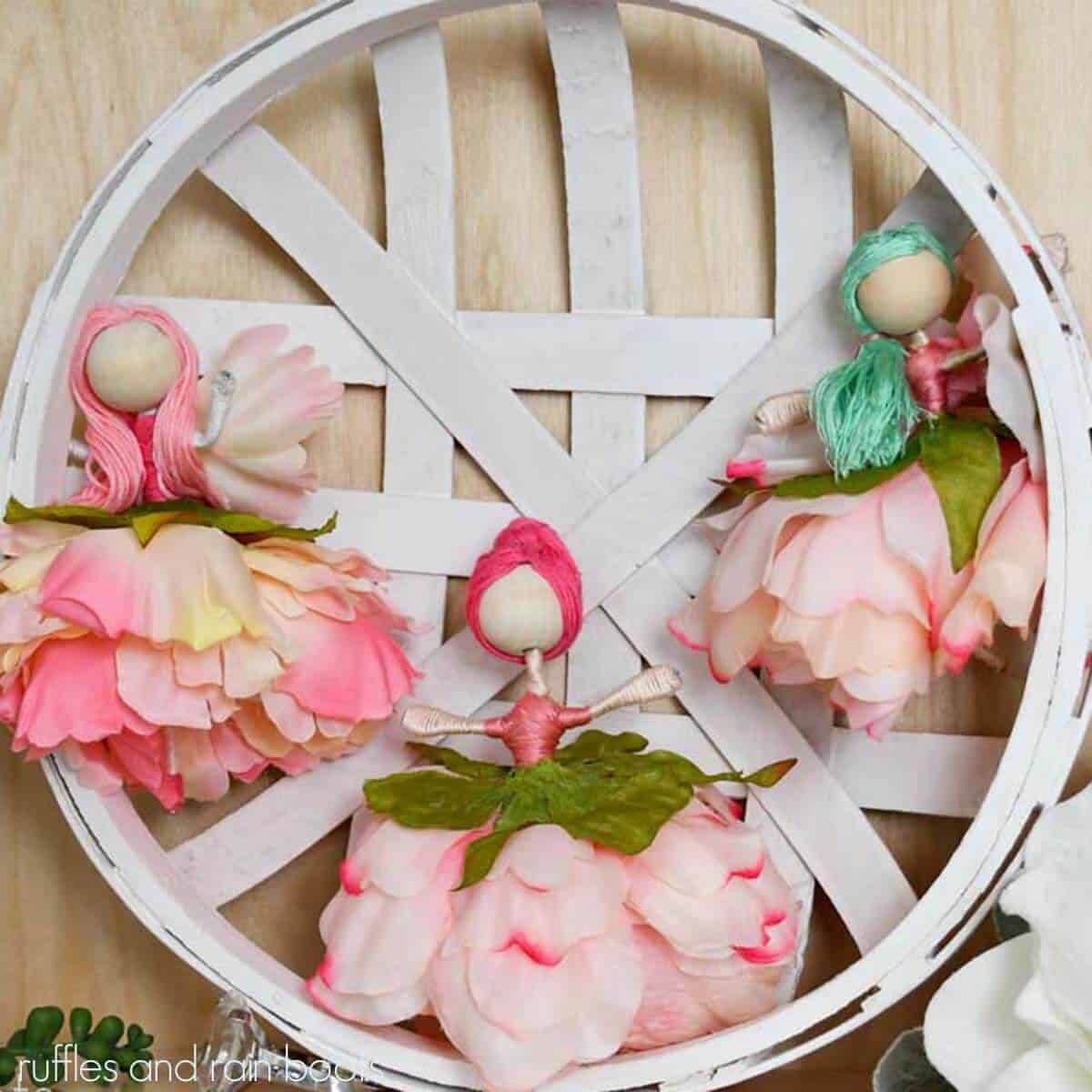 Square close up image of silk flower fairy dolls with wood bead heads on a white farmhouse basket.