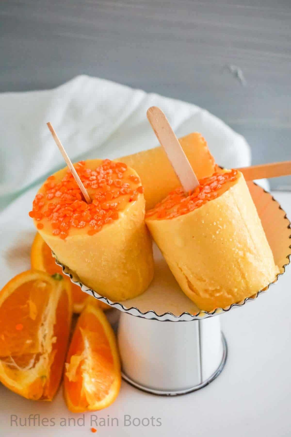 Vertical image of three orange Julius Dixie cup pops on a cake tray with orange slices and a white table.