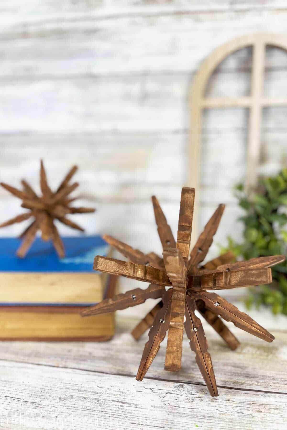 Vertical image close up of a brown wood clothespin star in 3D style in front of a farmhouse display and white washed wood background.