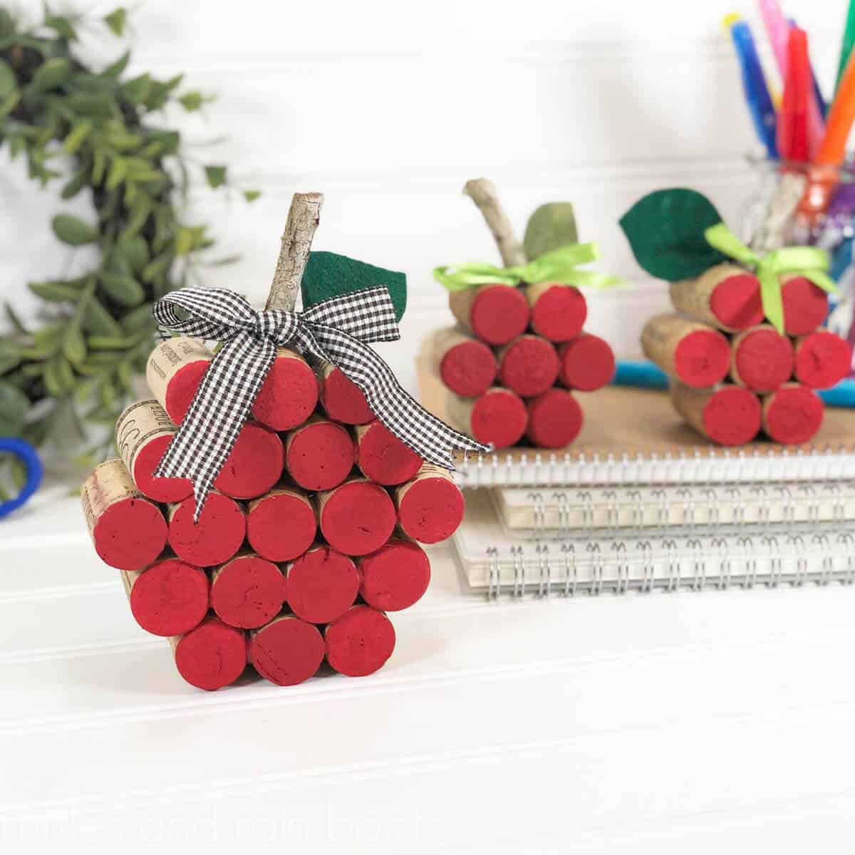 Square close up image of three wine cork apples painted red with leaves and ribbon in front of a wreath.