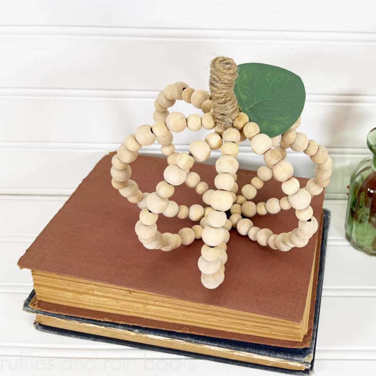 Square close up of a wood bead pumpkin with a twine stem and faux leaf sitting on a stack of books.
