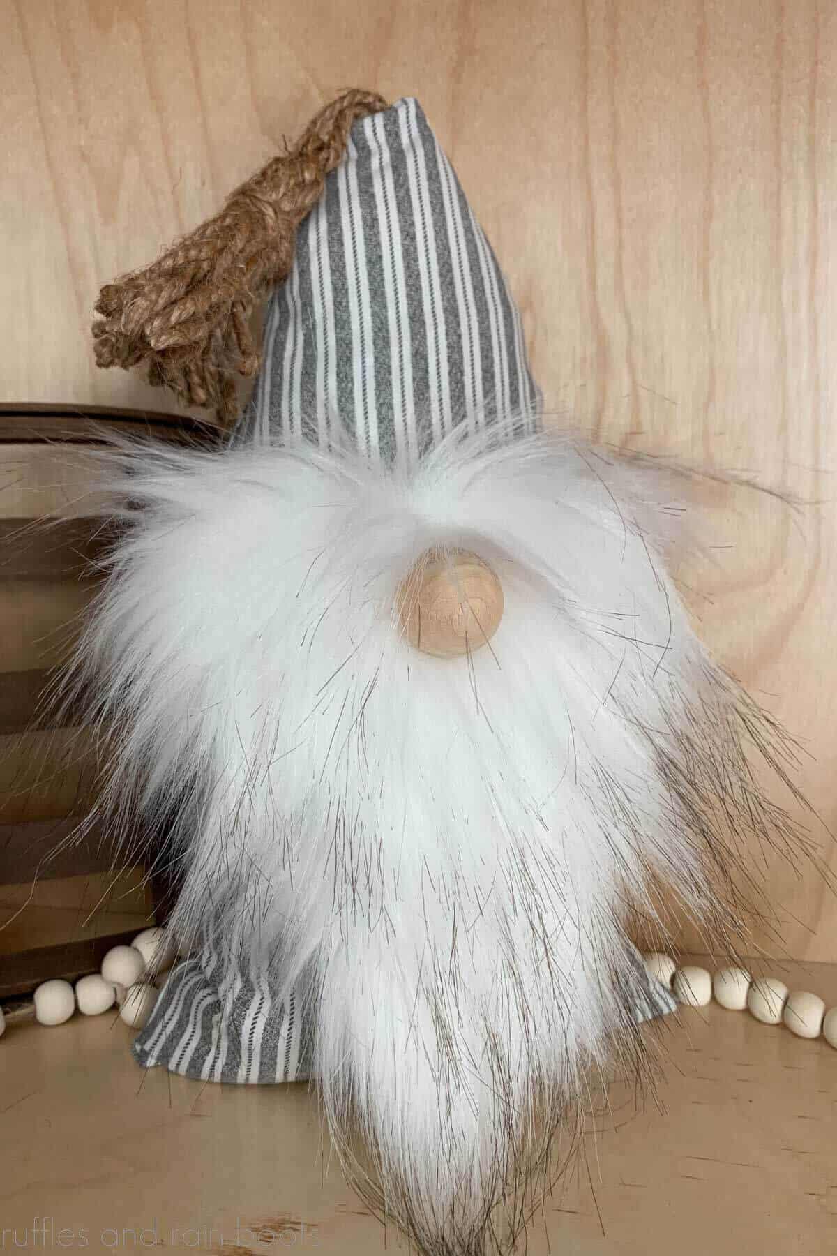No sew pillow gnome with a striped body and white beard against a light wood background with farmhouse beads.