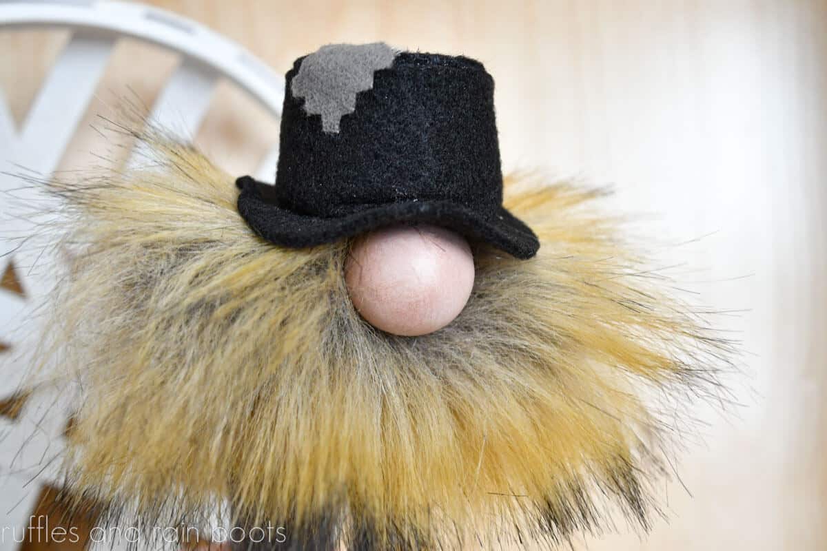 Horizontal close up image of a pompom gnome wearing a hat with patch in front of a light wood background.