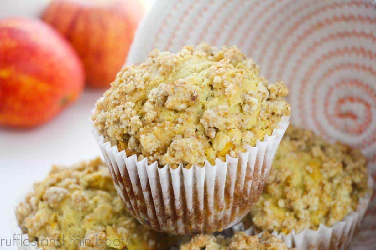 Horizontal image close up of stacked apple muffins next to a woven orange and white bowl and apples.