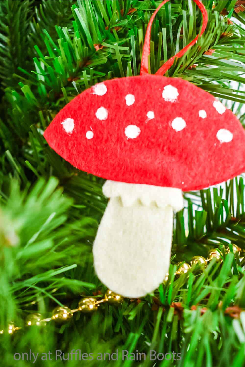 Vertical close up image of a red and white mushroom felt ornament hanging on a tree with a gold bead garland.