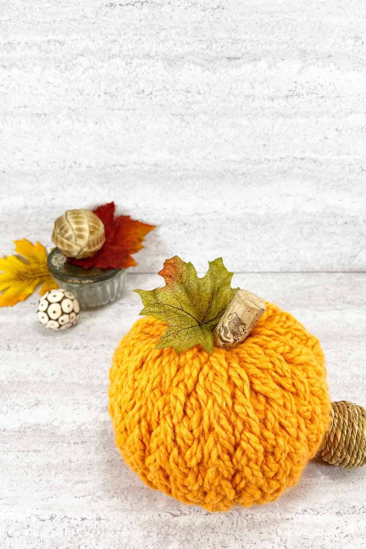 Vertical image of an orange sweater pumpkin made with yarn, fall leaves, and a wine cork against a concrete background.