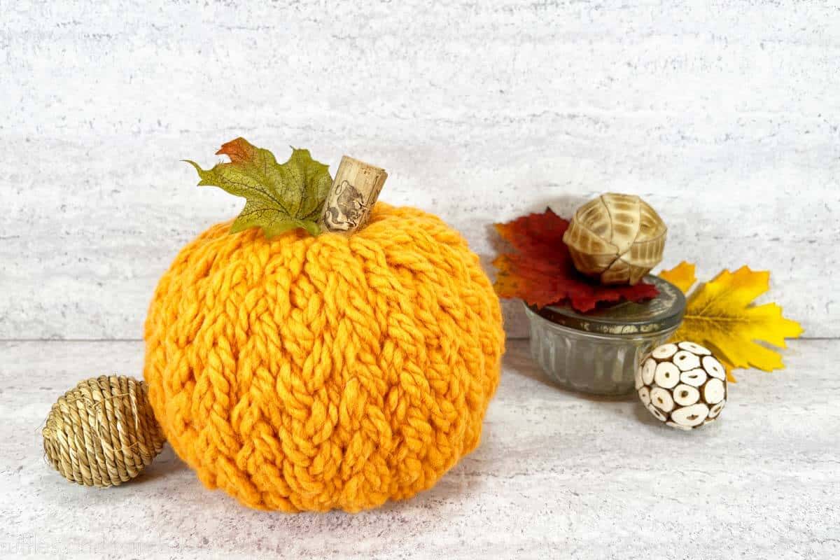 Horizontal image of a yarn-wrapped sweater pumpkin DIY with vase filler and fall leaves against a concrete background.
