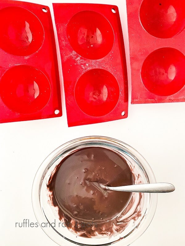 Melted chocolate in a glass bowl with spoon and red sphere mold on a white surface.