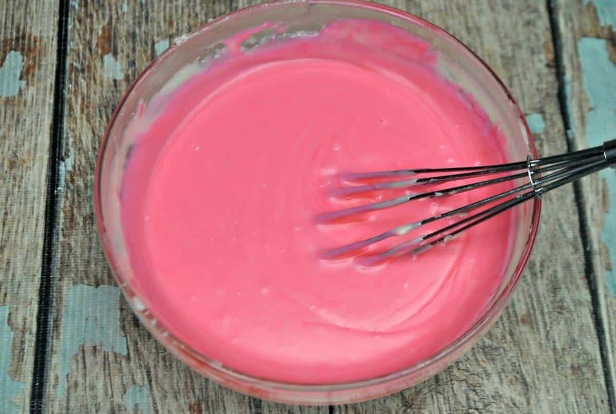 Overhead close up of melted pink fudge in a glass bowl with a whisk on a blue and gray weathered wood.