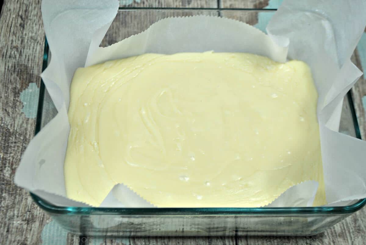 Overhead close up of melted white fudge mixture in square glass pan lined with parchment paper on a blue and gray weathered wood