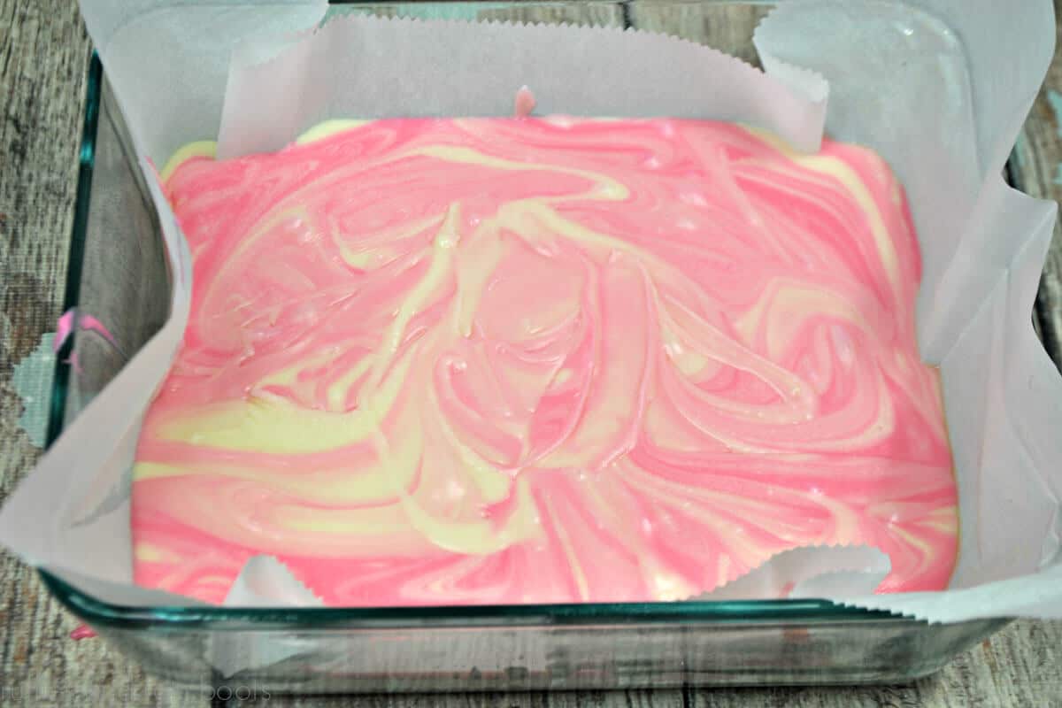 Overhead close up of swirled pink and white fudge mixture in square glass pan lined with parchment paper on a blue and gray weathered wood
