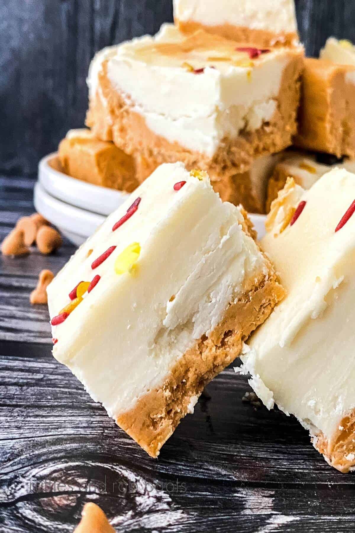 Close up of a Butterbeer Fudge Recipe square in front of white plates with additional Butterbeer Fudge on a dark gray weathered wood.