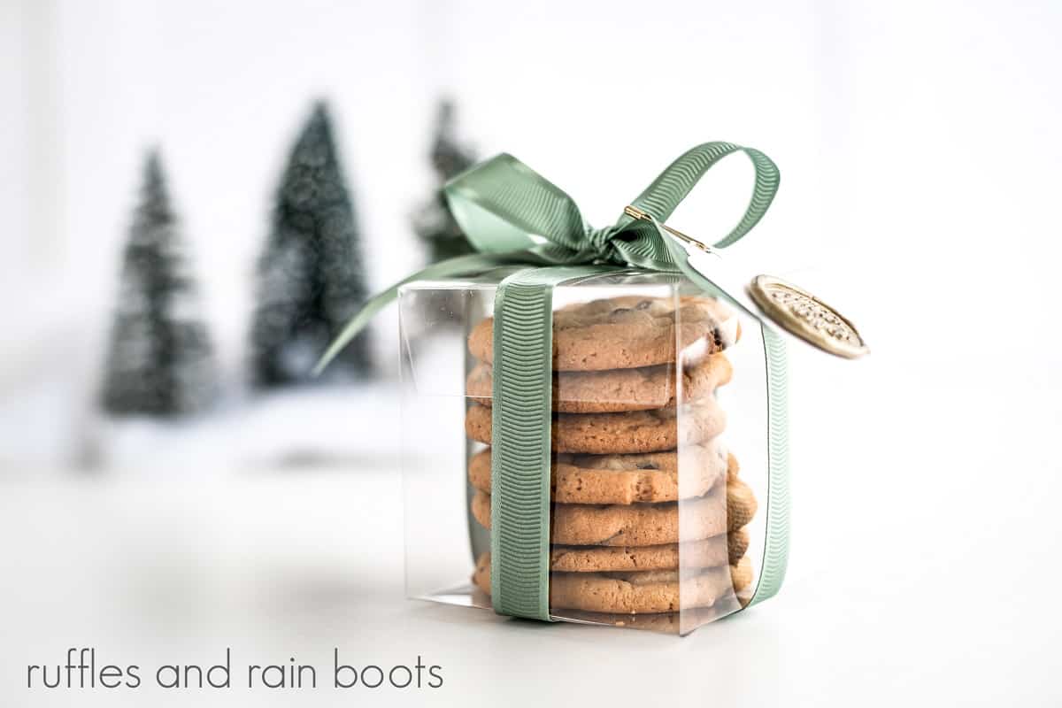 Close up side view of a Cricut Cookie Gift Box filled with chocolate chip cookies, wrapped and tied in ribbon with a homemade gift tag with a seal pinned to the ribbon with 3 mini Christmas trees in the background on a white surface.