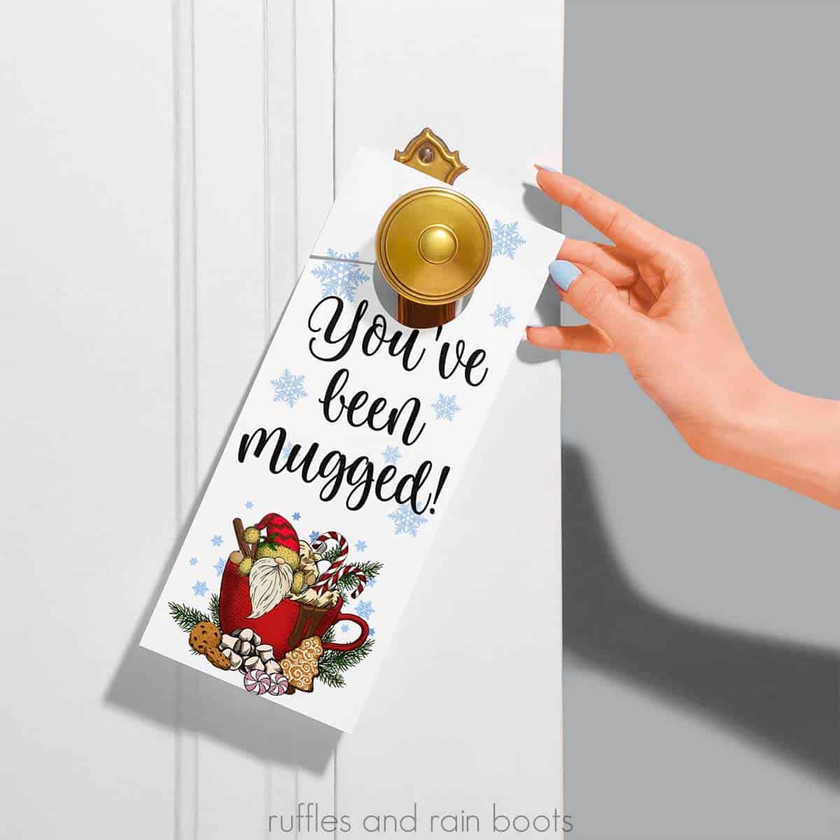 Square close up image of a You've been mugged door tag with a gnome and snowflakes on white door with gray wall and a hand.
