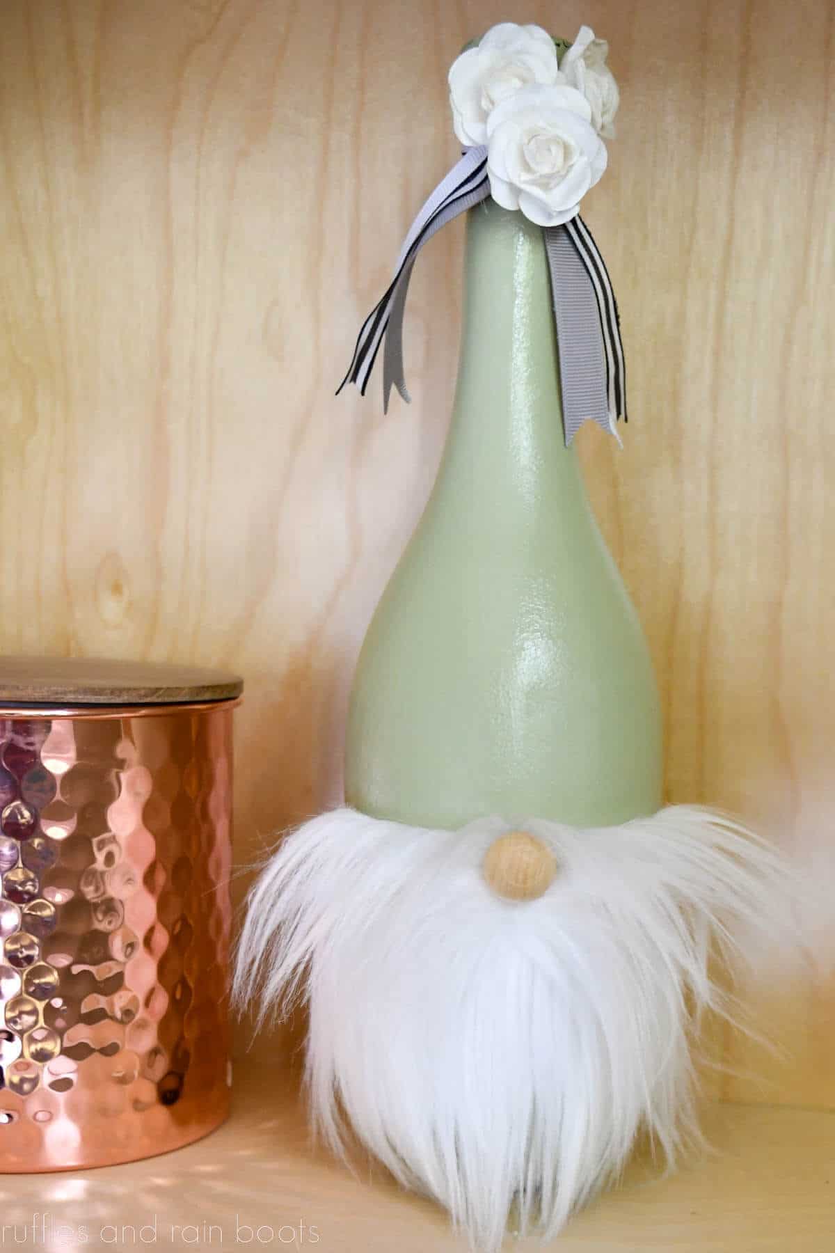 Vertical image of a painted wine bottle gnome with white beard, ribbon, and flower decorations on light wood background with copper vase.