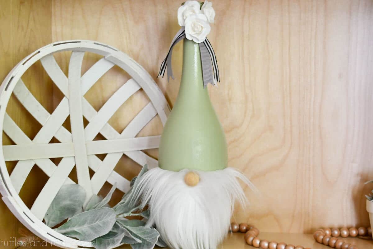 Horizontal close up image of a wine bottle gnome made with paint, fur, ribbon, and flowers placed on a light wood background.