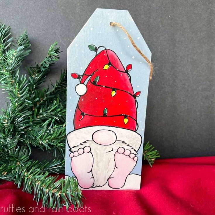 Square close up image of a painted Christmas gnome on a wood tag in front of a slate background on red fabric with pine accents.