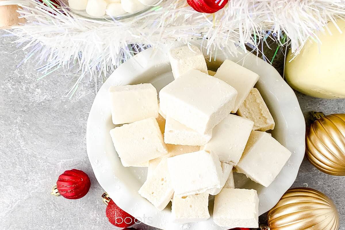 Overhead image of homemade eggnog marshmallows in a white ruffled bowl with Christmas decor in the back and red and gold Christmas ornaments in the front on a grey surface.