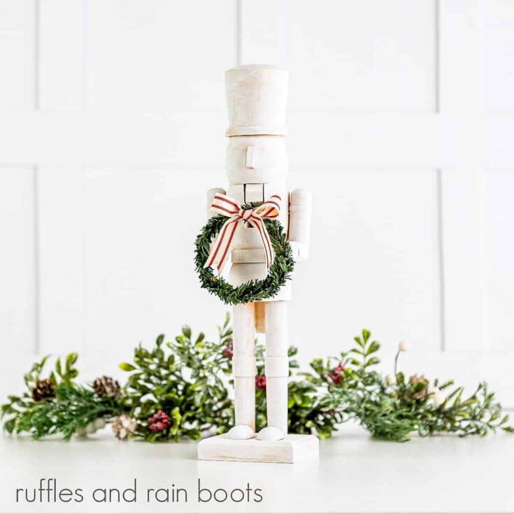 Front right facing view of a white painted wooden nutcracker holding a faux green wreath with a small red and white bow with faux greenery in the background on a white surface.