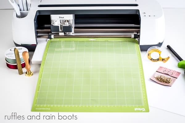 Cricut machine loaded with a green mat and acetate with tools, ribbon, a wax sealer set, safety pins, double sided tape and a lighter around it on a white surface.