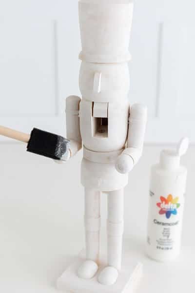 A white wooden nutcracker being painted white with a foam brush next to a bottle of white paint on a white surface.