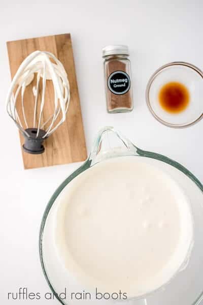 Overhead image of water and gelatin in a glass mixing bowl on a white surface next to a bottle of nutmeg, a bowl of vanilla and a whisk attachment on a small wooden board, all on a white surface.