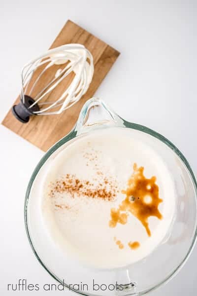 Overhead image of white gelatin mixture with nutmeg and vanilla on top in a glass mixing bowl on a white surface next to a whisk attachment on a small wooden board.