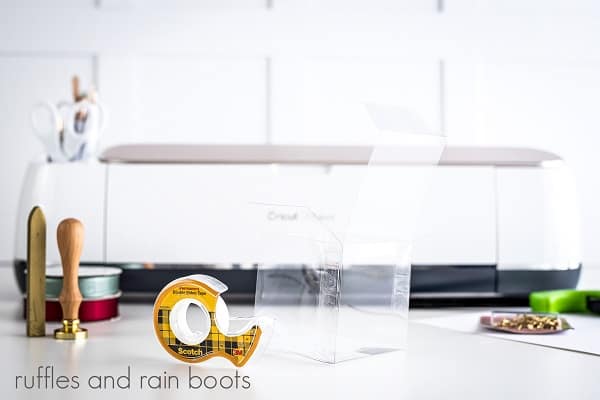 Cricut machine and tools in the background with folded cookie gift box surrounded by ribbon, safety pins, double sided tape and the seal kit on a white surface.