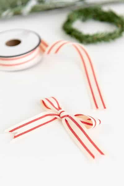 Close up of a small red and white bow next to a spool of white and red ribbon and a mini faux green wreath on a white surface.